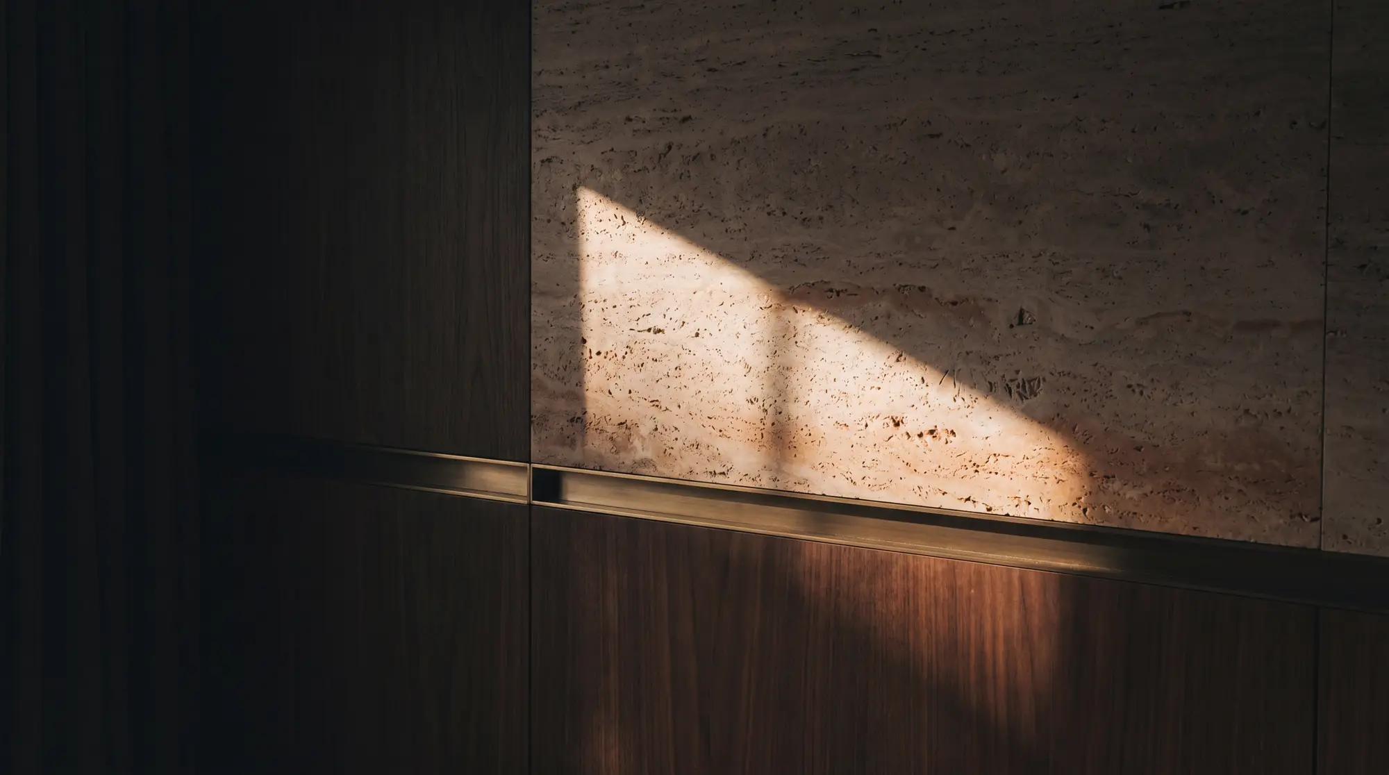 Detail of honed travertine meeting walnut with a brushed-brass reveal, raked by warm light